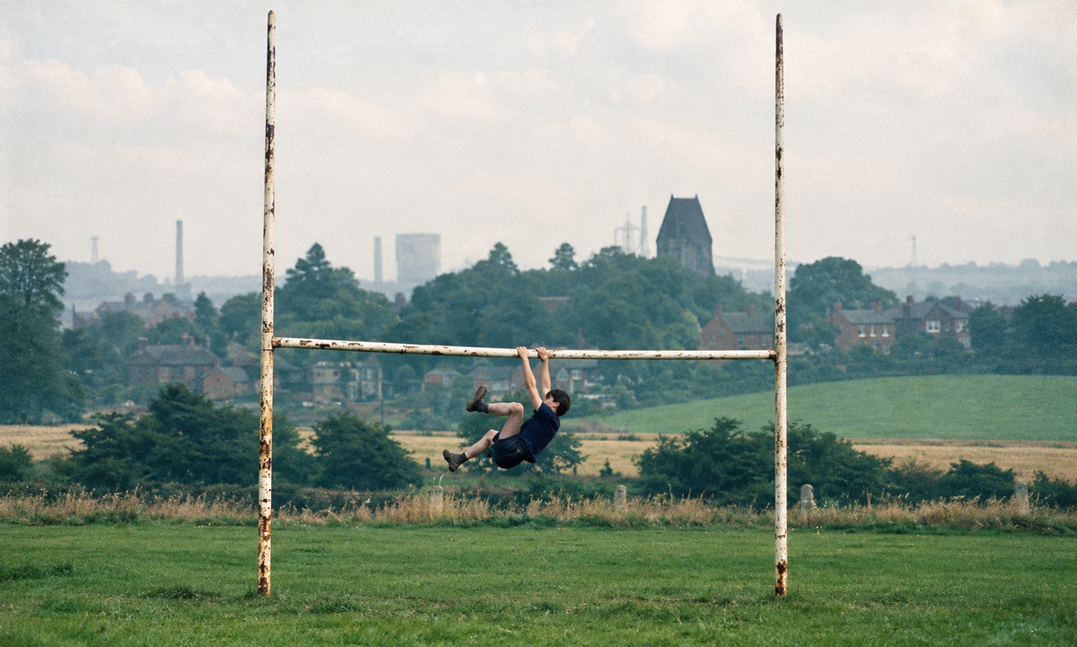 A boy hanging from the crossbar of old rugby posts on a local pitch